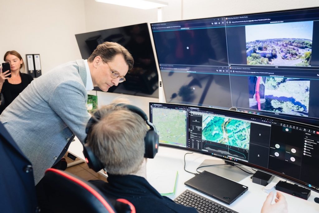 Minister President of NRW Hendrik Wüst stands in front of several computer screens together with a drone operator witnessing HHLA Sky's ICC